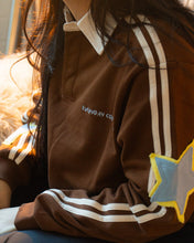 Load image into Gallery viewer, A close-up lifestyle shot of a person wearing a brown long-sleeve collared sweatshirt with white stripes on the sleeves, featuring a blue and yellow star patch on the sleeve and white embroidered text on the chest