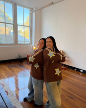 Load image into Gallery viewer, Two women standing together in a bright room with hardwood floors and large windows, wearing matching brown sweaters with yellow star patches and light blue jeans.