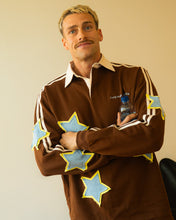 Load image into Gallery viewer, A man with a mustache wearing a brown long-sleeve rugby shirt with light blue and yellow star patches, posed against a plain beige wall.
