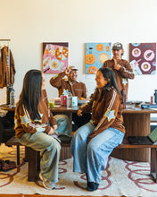 Load image into Gallery viewer, Four people in a casual indoor setting wearing matching brown hoodies with colorful star and stripe graphics. Two people are seated on wooden benches, and two are standing behind a table with drinks and artwork on the wall.
