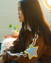 Load image into Gallery viewer, A woman in a brown sweatshirt with white stripes and a blue star patch sits in profile, illuminated by warm backlighting from a ring light. The sweatshirt has white stripes down the sleeves and a blue star patch outlined in yellow on the front.
