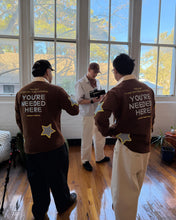 Load image into Gallery viewer, Three men standing in a bright room with large windows and hardwood floors, wearing matching brown shirts with 'YOU'RE NEEDED HERE' printed on the back in white text with a yellow star graphic.
